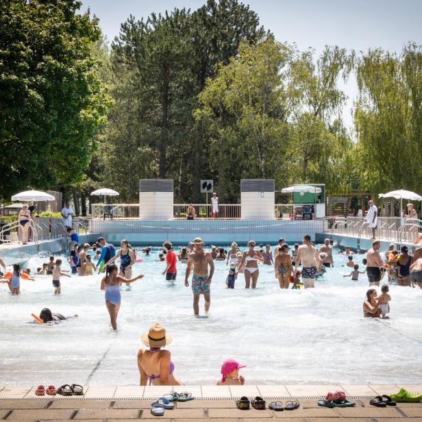 Viele Menschen, darunter Kinder und Erwachsene, genießen einen sonnigen Tag in einem Freibad mit flachem Wasser und umgebenden Bäumen.