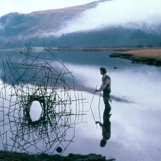 Ein Mann steht im Wasser neben einer Installation aus Ästen vor einer Bergkulisse.