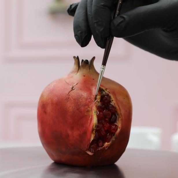 Eine Hand mit schwarzem Handschuh bemalt einen Granatapfel mit einem Pinsel.