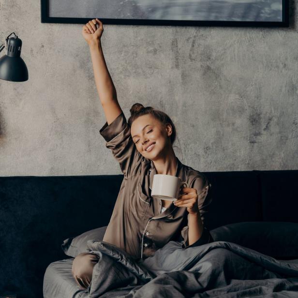 Eine Frau sitzt im Bett, streckt sich und hält eine Tasse in der Hand.