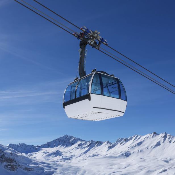 Eine Gondel schwebt vor schneebedeckten Bergen unter einem strahlend blauen Himmel.