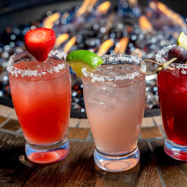 Drei farbenfrohe Cocktails mit Salzrand stehen vor einem Feuer.
