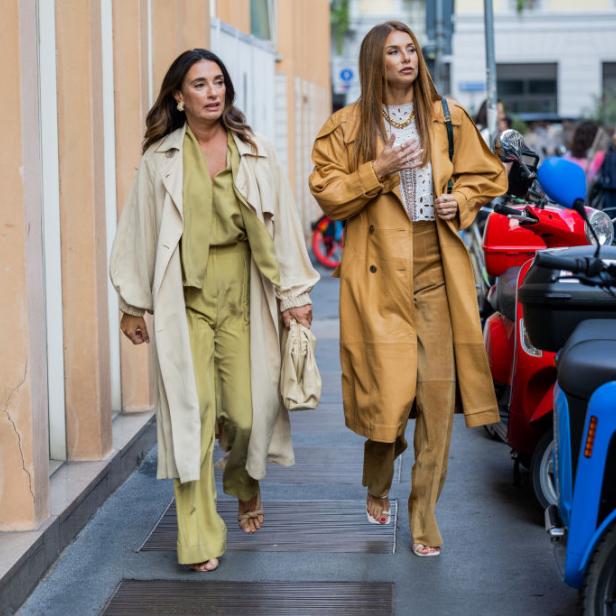 Zwei Frauen in Trenchcoats und passenden Hosen laufen auf einer Straße entlang.