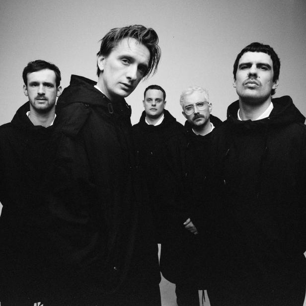 Schwarzweißaufnahme einer Band, die schwarze Kapuzenpullover vor einem neutralen Hintergrund trägt.