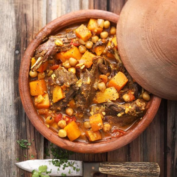 Ein Tajine-Gericht mit Fleisch, Kürbis und Kichererbsen in einem Tontopf auf einem Holztisch.