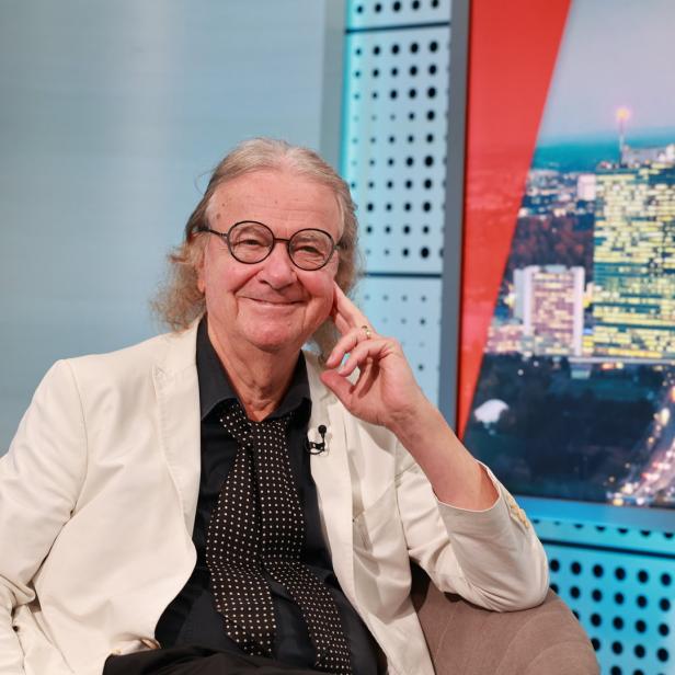Ein Mann mit Brille und weißem Sakko sitzt vor einem Hintergrund mit Stadtansicht.