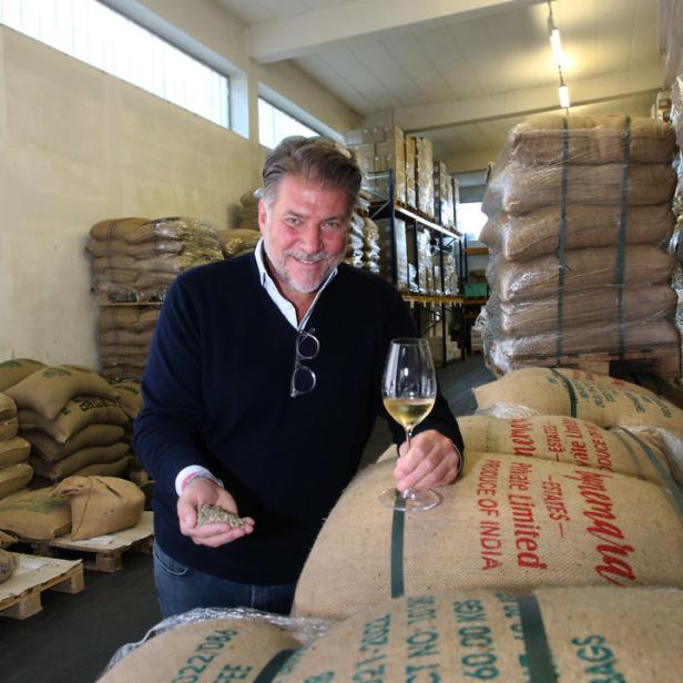 Ein Mann steht in einem Lagerhaus voller Kaffeesäcke und hält ein Glas Wein und Kaffeebohnen in der Hand.