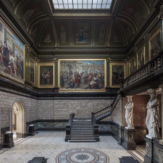 Ein weitläufiger Museumssaal mit Gemälden, Statuen und einer kunstvollen Treppe.