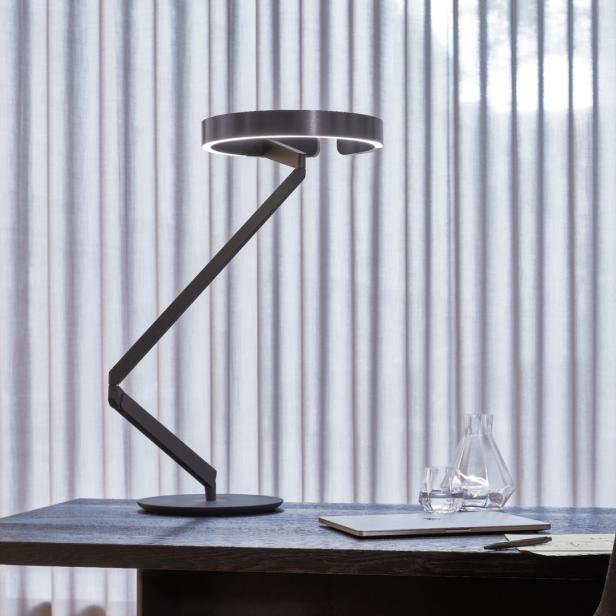 Eine moderne Schreibtischlampe beleuchtet einen Laptop und eine Wasserflasche auf einem dunklen Tisch.