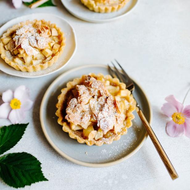 Drei Apfeltörtchen mit Mandeln und Puderzucker auf Tellern mit Blumen dekoriert.