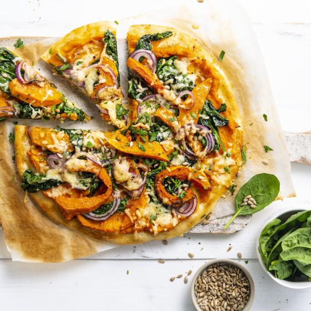 Eine Pizza mit Kürbis, Spinat, roten Zwiebeln und Käse auf einem Holztisch.