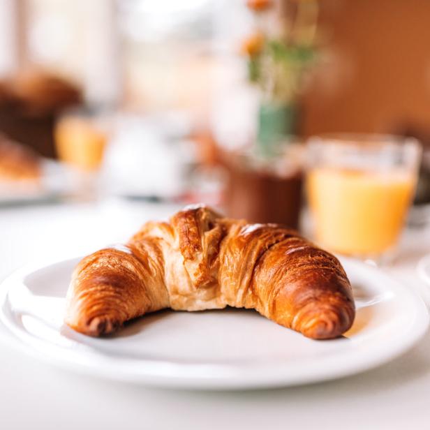 Ein Croissant liegt auf einem Teller, zusammen mit Orangensaft und Marmelade zum Frühstück.