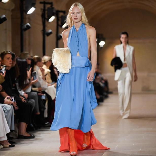 Eine blonde Frau in einem blauen Kleid und orangefarbenen Stiefeln läuft über einen Laufsteg.