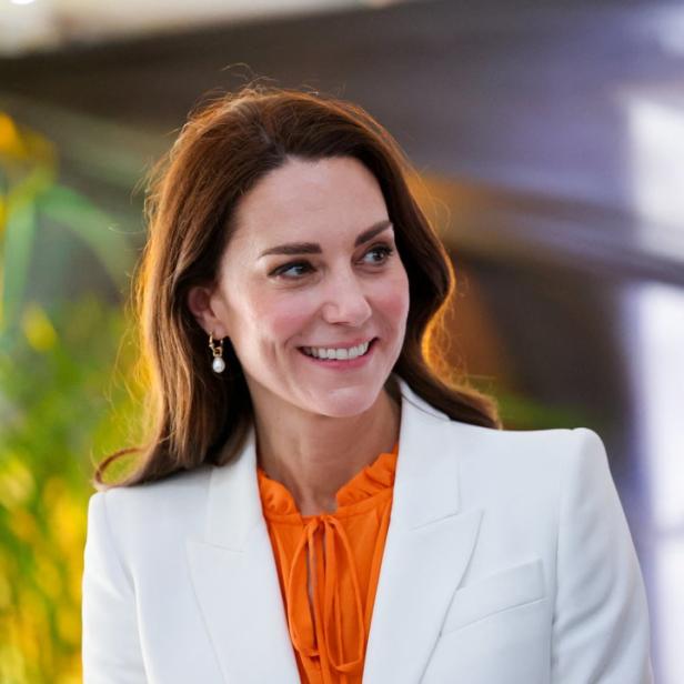 Kate Middleton lächelt in einem weißen Blazer und einer orangefarbenen Bluse.