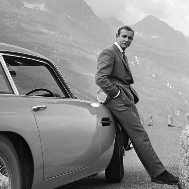 Sean Connery lehnt an einem Aston Martin vor einer Bergkulisse.