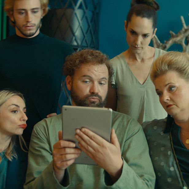 Eine Gruppe von fünf Personen schaut aufmerksam auf ein Tablet in einem Wohnzimmer.