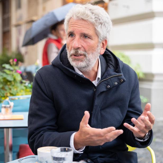 Ein Mann mit grauem Haar gestikuliert an einem Tisch im Freien.