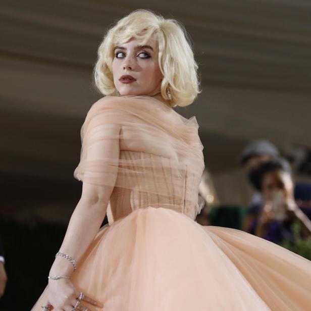 Billie Eilish in einem langen, pfirsichfarbenen Kleid mit blonden Haaren, die über die Schulter blickt.