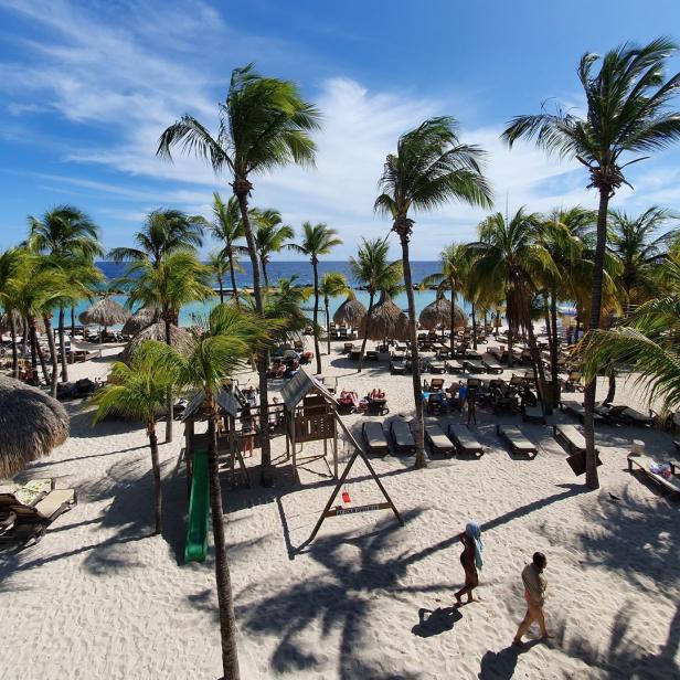 Ein Sandstrand mit Palmen, Liegestühlen und einigen Personen unter blauem Himmel.