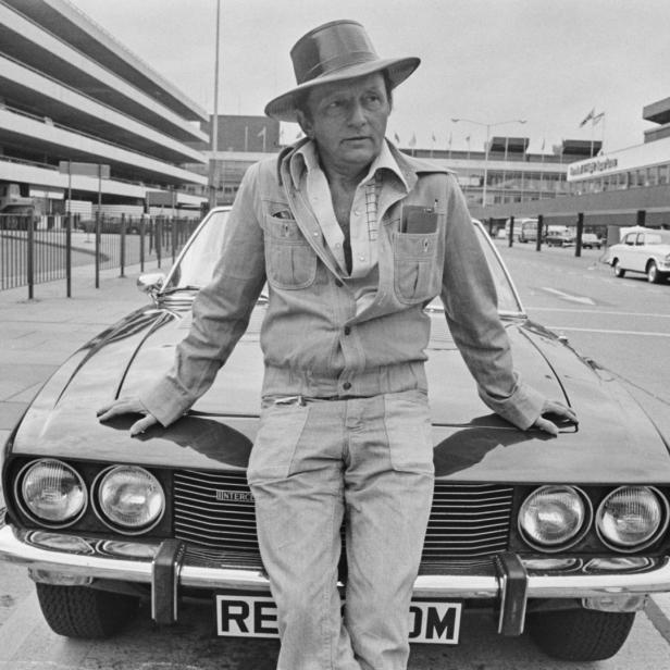 Ein Mann mit Hut lehnt an einem Jensen Interceptor vor einem Flughafengebäude.