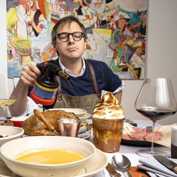 Ein Mann mit Brille flambiert ein Dessert mit einem Brenner an einem reich gedeckten Tisch.