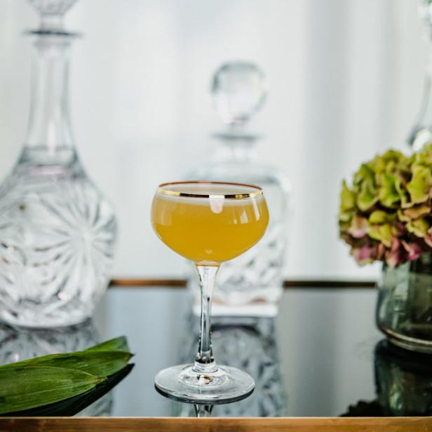 Ein Cocktail mit goldener Umrandung steht auf einem Tisch mit Karaffen und Blumen.