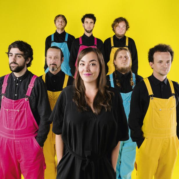 Eine Gruppe von acht Personen vor einem gelben Hintergrund, gekleidet in farbenfrohe Overalls.