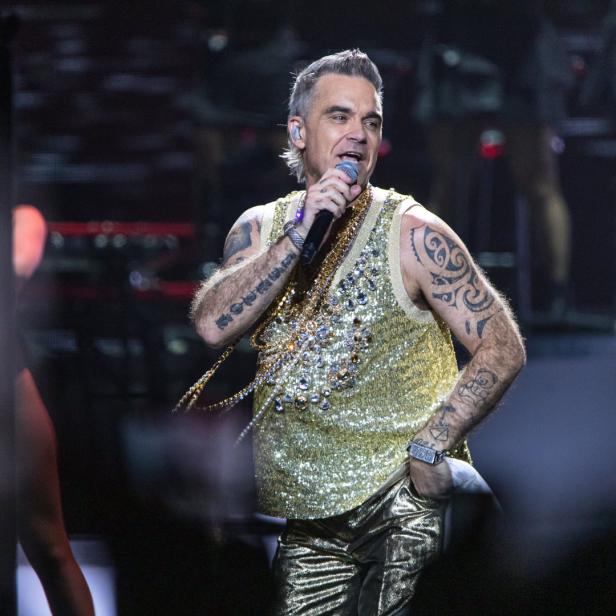 Robbie Williams tritt in einem goldenen Glitzertop und goldenen Hosen auf der Bühne auf.