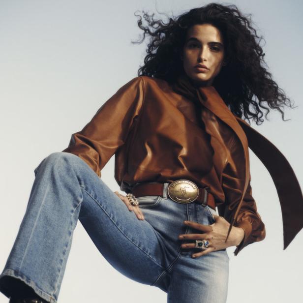 Eine Frau mit lockigem Haar posiert in Jeans, Lederbluse und Cowboystiefeln.