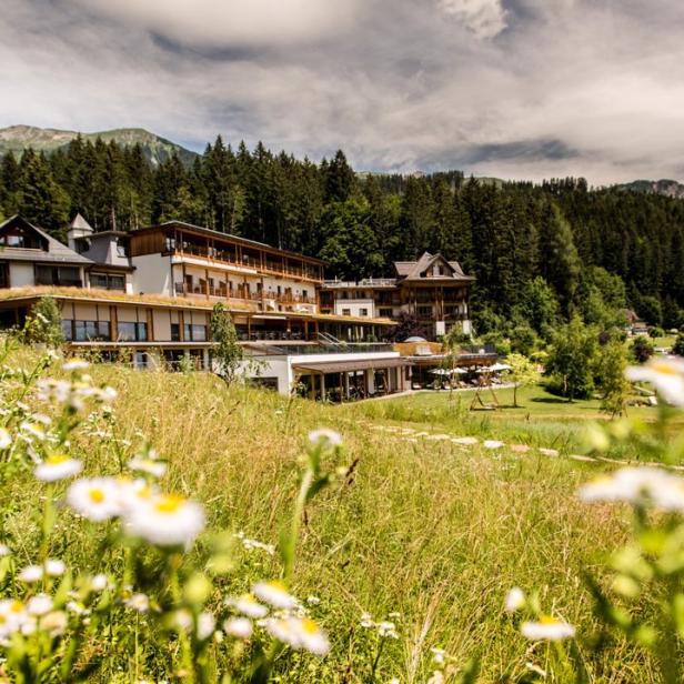 Ein Hotel liegt eingebettet in eine grüne Landschaft mit Wiesen und Bergen im Hintergrund.