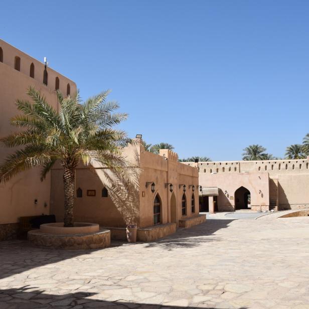 Ein Innenhof einer historischen Festung mit einer Palme und sandfarbenen Gebäuden unter blauem Himmel.