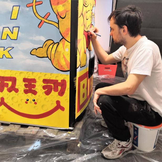 Ein Mann bemalt eine bunte Werbetafel mit der Aufschrift „INK“ und japanischen Schriftzeichen.