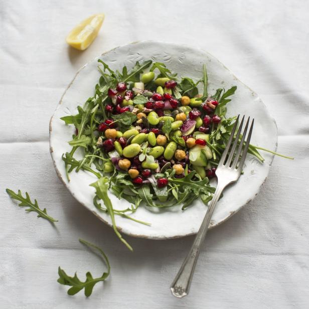 Ein Teller Salat mit Rucola, Edamame, Kichererbsen und Granatapfelkernen steht auf einer weißen Tischdecke.