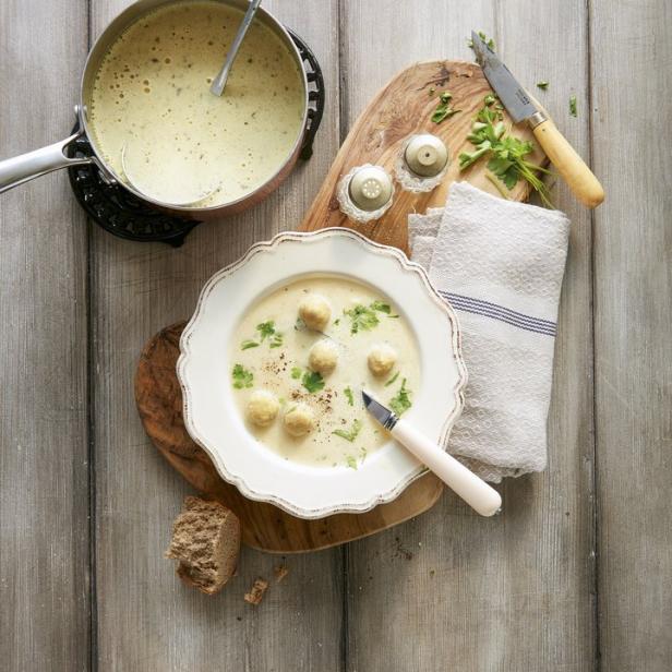 Eine Schüssel mit Suppe und Klößchen, garniert mit Petersilie, neben einem Topf und Brot auf einem Holztisch.