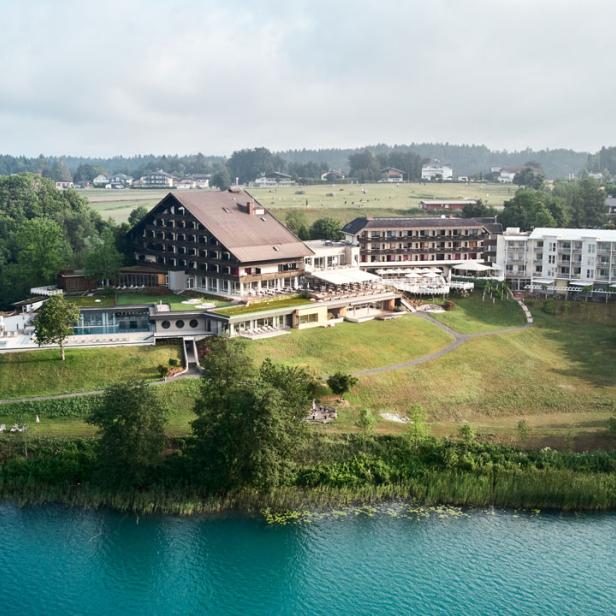 Ein Hotelresort liegt an einem Seeufer mit türkisfarbenem Wasser.
