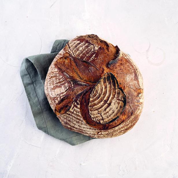 Ein Laib Sauerteigbrot liegt auf einem grauen Tuch auf einer weißen Oberfläche.