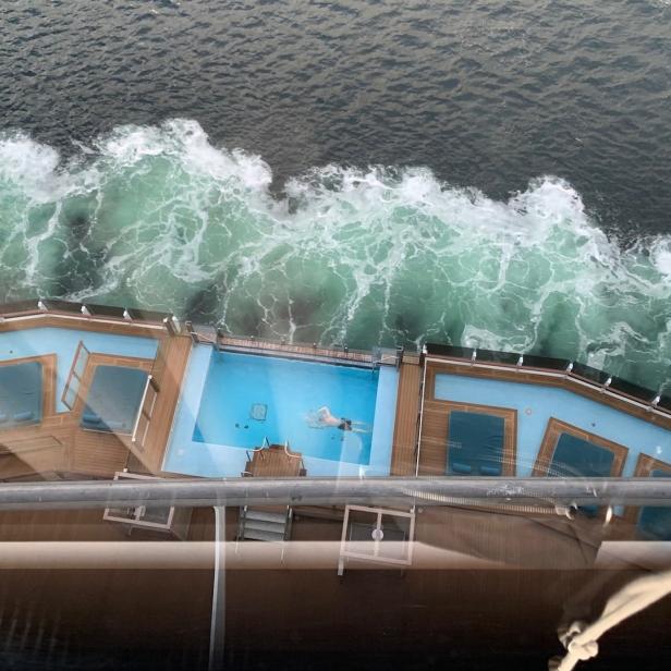 Eine Person schwimmt im Pool eines Kreuzfahrtschiffes, während Wellen daneben ans Ufer schlagen.