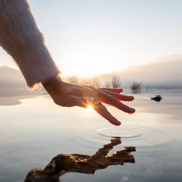 Eine Hand berührt die Wasseroberfläche eines Sees bei Sonnenaufgang.