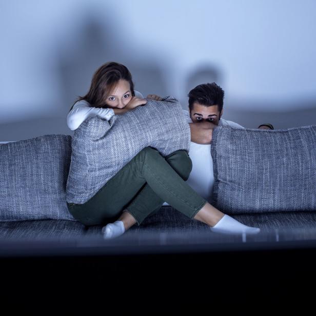 Ein Paar versteckt sich ängstlich hinter Kissen auf einem Sofa.