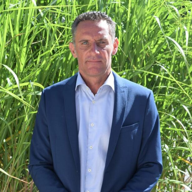 Ein Mann im blauen Anzug steht vor hohen grünen Gräsern und blickt ernst in die Kamera.