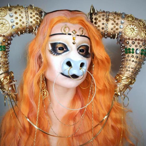 Eine Person mit orangefarbenen Haaren und aufwendigem Make-up, das an eine mythologische Kreatur erinnert, posiert.