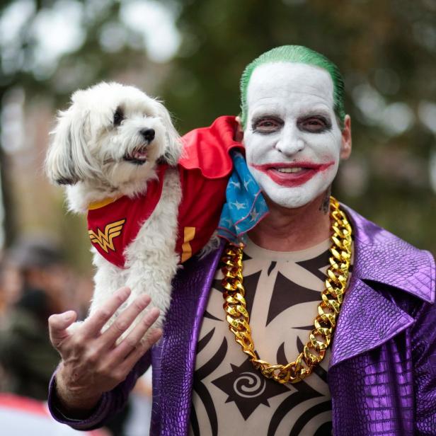 Ein Mann, als Joker verkleidet, trägt einen Hund im Wonder-Woman-Kostüm auf der Schulter.