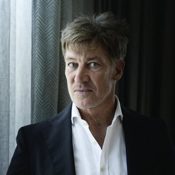 Ein Mann mit blau-grauen Augen und grau meliertem Haar trägt ein weißes Hemd und einen schwarzen Blazer.