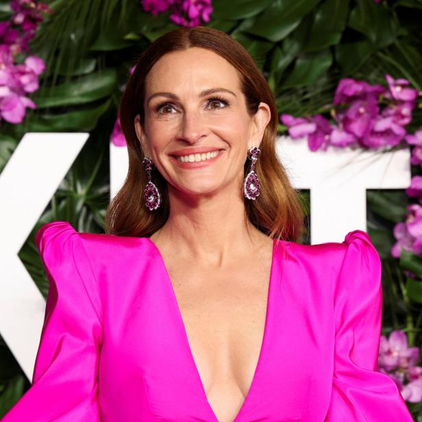 Julia Roberts lächelt in einem pinkfarbenen Kleid vor einem floralen Hintergrund.