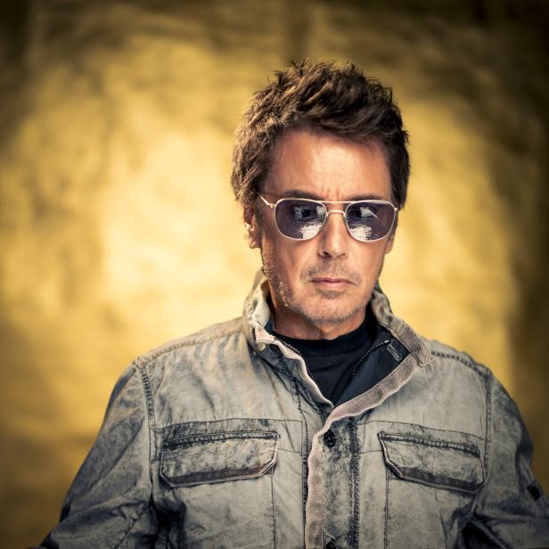 Ein Mann mit Sonnenbrille und grauer Jacke vor einem goldenen Hintergrund.