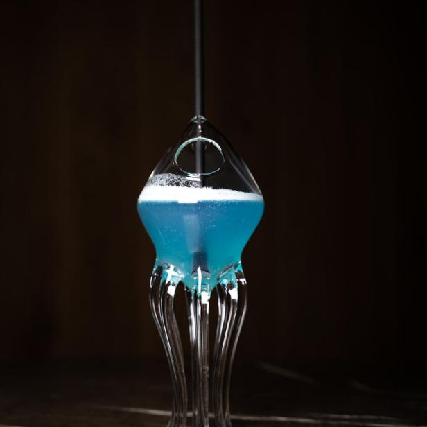 Ein blauer Cocktail in einem ungewöhnlichen Glas mit einem Strohhalm steht auf einem Tisch.