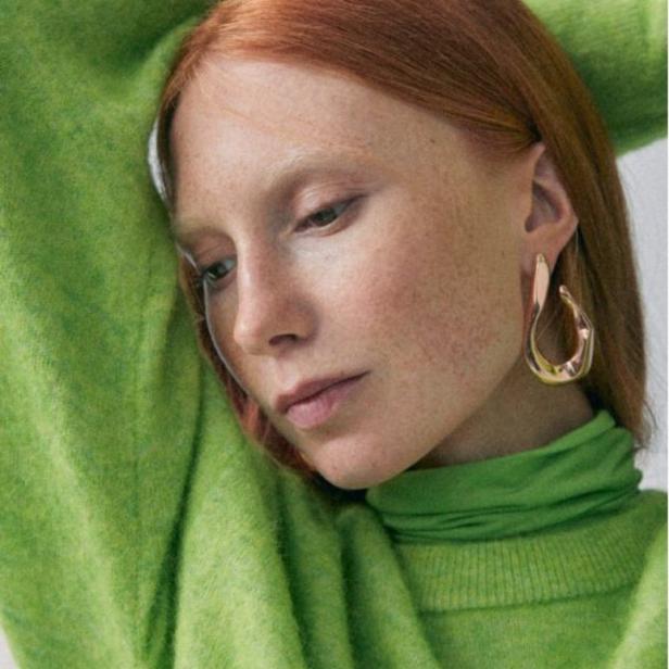 Eine Frau mit roten Haaren und Sommersprossen trägt einen grünen Rollkragenpullover und goldene Ohrringe.