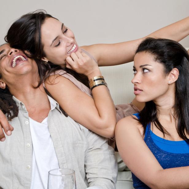 Drei junge Erwachsene sitzen zusammen auf einem Sofa, wobei eine Frau eifersüchtig wirkt, während die anderen beiden lachen.