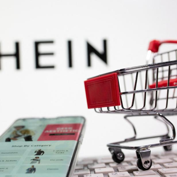 Ein Mini-Einkaufswagen steht auf einer Tastatur, im Hintergrund das SHEIN-Logo und ein Smartphone mit der SHEIN-App.