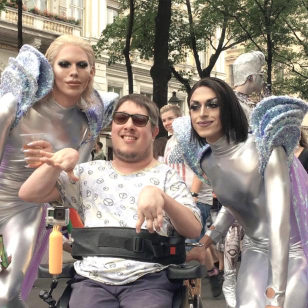 Ein Mann im Rollstuhl posiert mit zwei Dragqueens in silbernen Kostümen für ein Foto.